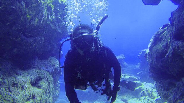selfie camera view of a scuba diver wearing diving gear swimming through a under water narrow small cave gap between coral reef rocks toward other divers in front in blue ocean sea bottom during day