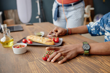Young man arranging cherry tomatoes on wooden table near long baguette and small bowls of olive oil and salsa during food photography session