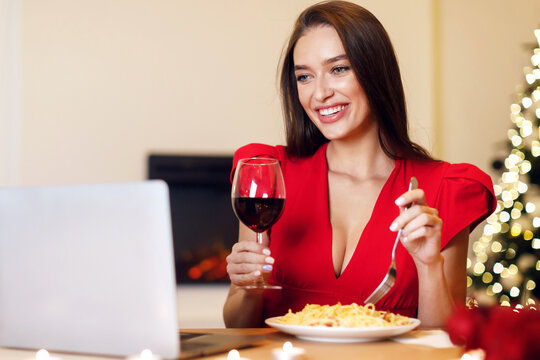 Virtual Party. Happy beautiful woman making videocall with friends, boyfriend or husband during dinner, sitting at dining table eating pasta and drinking red wine, looking at screen, celebrating alone