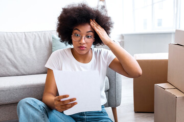 Large Expenses. Shocked Black Lady Reading Bill Sitting Among Moving Boxes In New Home. Renting Apartment Prices, Mortgage and Real Estate Problem, House Ownership Papers Issue Concept