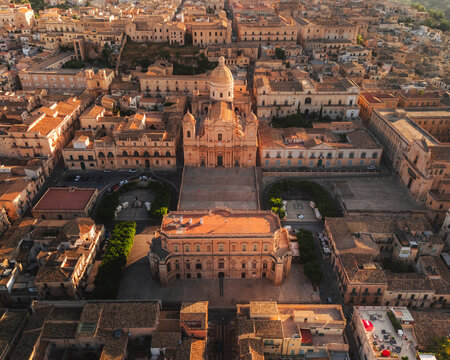 Aerial view of the golden light bathing the Baroque architecture of Noto Cathedral and surrounding buildings, casting long shadows across the city's intricate street pattern, Noto, Sicily, Italy.