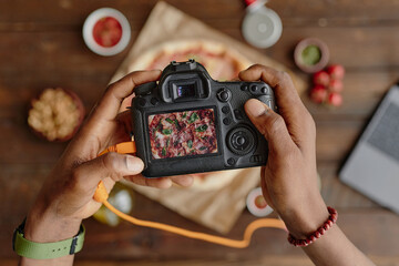 Man photographing pizza with professional camera, hands holding camera above pizza on wooden table, capturing food image for culinary project, laptop and ingredients visible