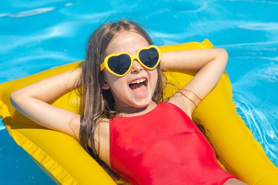Close up portrait little girl in red swimsuit and yellow heart-shaped sunglasses lies on inflatable mattress in swimming pool with blue water on warm summer day on tropical vacation. Childhood concept
