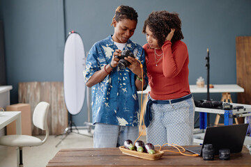 Young adult Black man and young adult Black woman reviewing photos on digital camera in professional food photography studio, standing near table with vegetables and photography equipment