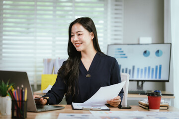 A smiling young woman reviews documents at her desk, with a laptop, calculator, and graphs on a...