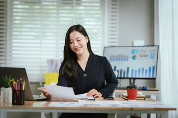 A smiling young woman reviews documents at her desk, with a laptop, calculator, and graphs on a...