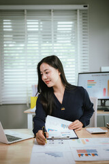 A smiling young woman reviews documents at her desk, with a laptop, calculator, and graphs on a...
