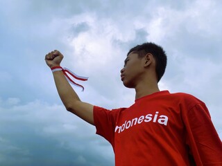 Indonesian boy with a red and white ribbon clenching his fist towards a blue-grey sky during Indonesia's Independence Day celebration