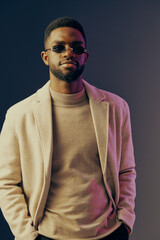 Stylish young African American man in sunglasses wearing a beige blazer, posing confidently against a dark colorful background, showcasing modern fashion trends