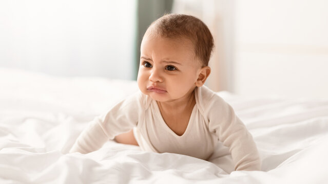 Portrait of crying African American infant in bodysuit whining, crawling on white bedsheets in bedroom at home indoors. Closeup of irritated baby feeling upset, banner, blurred background
