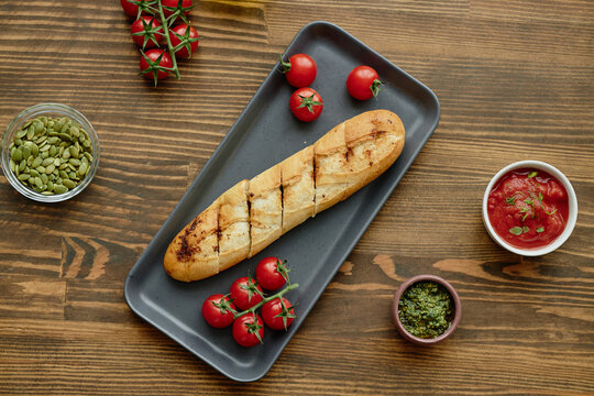 Grilled baguette slices arranged on rectangular plate surrounded by fresh cherry tomatoes, bowl of tomato sauce, small bowl of green pesto, glass bowl of pumpkin seeds on wooden table - Powered by Adobe