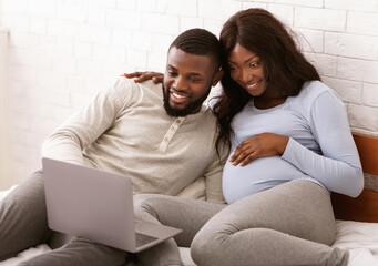 Happy pregnant african couple choosing furniture for baby room online, using laptop, laying on bed, empty space