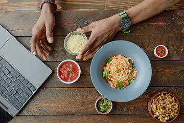 Young man reaching for bowl of grated cheese while sitting at wooden table with plate of pasta, tomato sauce, pesto, walnuts and open laptop visible in workspace