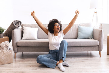 Fototapeta premium Happy African American Woman Sitting Among Moving Boxes Indoor, Shaking Fists Celebrating Relocation To New Home. Real Estate, Renting Apartment Service, Housing Concept
