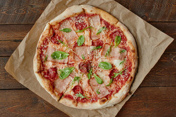 Whole freshly baked pizza with slices of ham, pepperoni, tomato sauce, grated cheese and green basil leaves arranged on brown parchment paper over rustic wooden table
