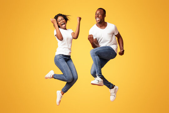 Emotional african american young couple winners clenching fists and screaming, jumping up over orange studio background. Happy black man and woman celebrating success, full length photo