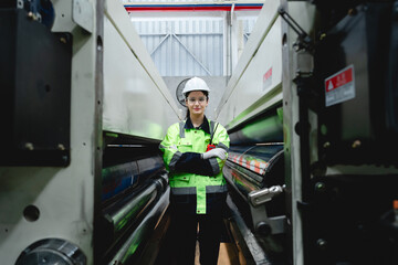 Naklejka premium Confident female printing factory worker wearing safety gear standing between two large printing machines with walkie-talkie in hand.