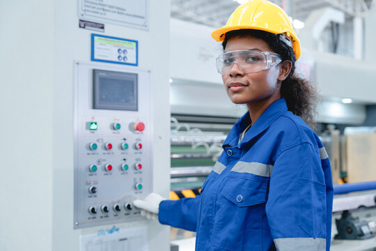 Portrait of Hispanic female technician in safety gear operating control panel at modern manufacturing plant, smiling confidently at camera.
