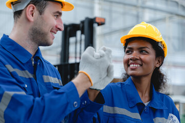 Male and female industrial engineers in blue uniforms and safety helmets smiling and giving a high five, showing team spirit and cooperation in factory workplace.