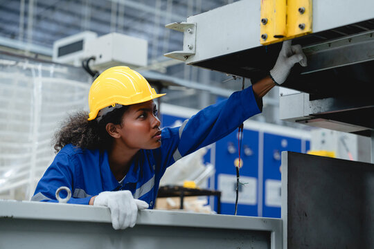 Female industrial worker wearing a yellow helmet and blue uniform examines a cardboard processing machine in a modern paper box factory, showing precision and safety in her task.
 - Powered by Adobe