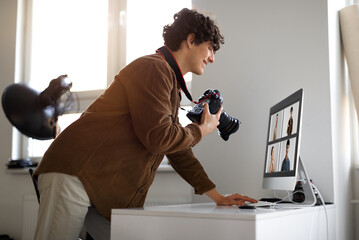 Creative job. Happy young man photographer in casual outfit working at photo studio office, using digital camera and pc, checking photo shoot, side view