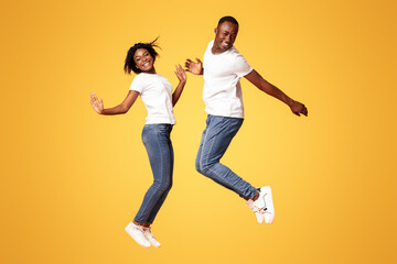 Cute and cheerful young black couple jumping up on orange studio background, full length shot, copy space. Happy african american lovers man and woman feeling joyful and free