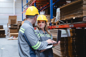 Professional logistics team checking cardboard supply data using digital tools and documents in storage facility with high stacked packaging boxes.
