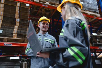 Male and female warehouse workers wearing safety helmets collaborate using laptop and clipboard while managing cardboard packaging inventory in modern factory.