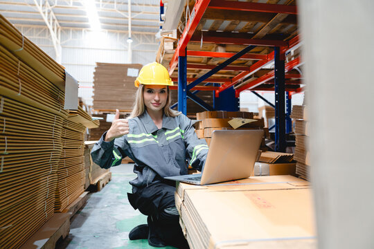 Confident female warehouse worker in safety uniform using laptop for inventory check in cardboard packaging area. Thumbs-up gesture shows successful operation in industrial storage zone. - Powered by Adobe