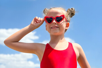 Cute little girl wearing red shirt top and heart-shaped sunglasses against a background of blue sky with white clouds. Carefree childhood, peace, love and freedom concept. High quality photo
