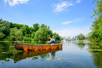healthy active lifestyle: young adult woman solo paddling canoe on a channel in the toronto islands with lush summer vegetation and city skyline in background room for text copy space