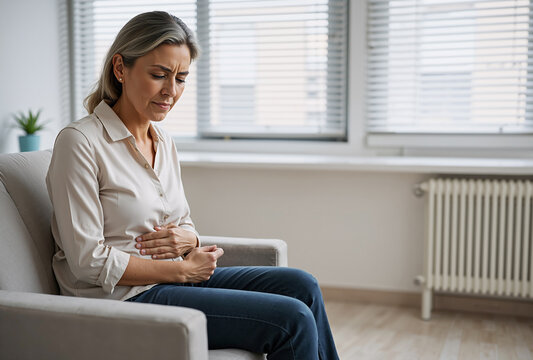 Middle-aged caucasian woman experiencing stomach pain while sitting on a beige couch in a bright living room with light decor and a somber mood, seeking comfort