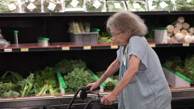 An elderly woman maneuvers her walker through a grocery store, selecting fresh vegetables while shopping for her weekly supplies.