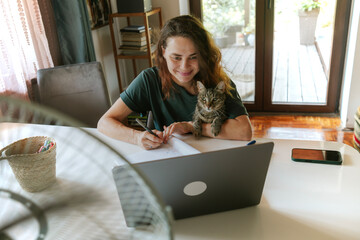 Young woman working at home remotely using a laptop with a grey cat on her lap