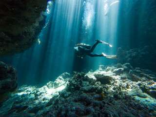 Landscape view of sunlight sunbeams dancing from the surface in underwater sea cave with rock walls and scuba diver exploring