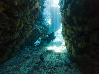 Landscape view of sunlight sunbeams dancing from the surface in underwater sea cave with rock walls and scuba diver exploring