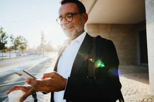 Senior businessman enjoying coffee while using smartphone outdoors in the morning - Powered by Adobe