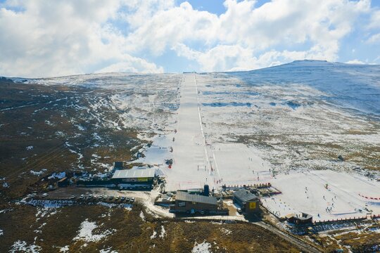 Aerial view of the snow-blanketed slopes of Afriski Mountain Resort, a stark white stripe against the brown landscape, dotted with skiers and vibrant flags, Butha-Buthe, Lesotho.