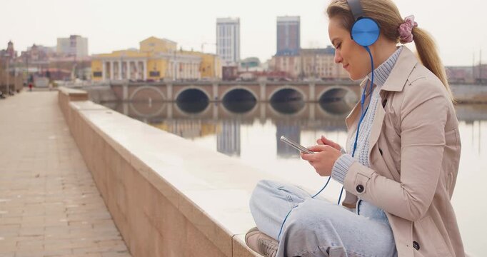 Slow motion zoom out footage of a 35 years old woman in headphones and listens to relaxing music and singing outdoor. Rest from office bustle and city noise. 4k footage.