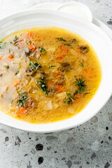 Bowl of clear vegetable soup with carrots, onion, and dill, served with sour cream on a white plate over gray speckled background. Top view, homemade comfort food.