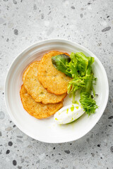 Three crispy potato pancakes served on a white plate with fresh salad and sour cream, garnished with olive oil on a speckled countertop. Top view.
