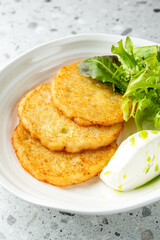 Three crispy potato pancakes served on a white plate with fresh salad and sour cream, garnished with olive oil on a speckled countertop. Top view.