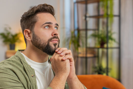 Tensed young man sitting on sofa at home folding palms together pray ask for help, thinks feel stressed looks desperate by life problems. Caucasian guy praying asking for help sitting on sofa at home.