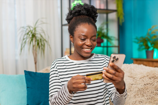 African American Black woman shopping online at home on sofa. Young girl entering credit card details on smartphone, smiling with joy, sitting in cozy living room. Technology, lifestyle, satisfaction