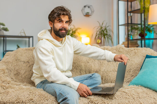 Young Indian man freelancer at home living room sitting on couch, opens laptop computer start working. Bearded guy works on notebook, sends messages, makes online purchases, watching movies, working.