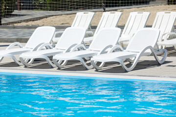 Empty poolside with white sun loungers and blue water. Row of white plastic sunbeds next to swimming pool on hotel outdoor patio. Summer vacation concept