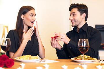 Proposal Date Concept. Smiling boyfriend holding open box, showing engagement ring to excited surprised girlfriend, proposing in luxury restaurant or at home. Couple having romantic dinner