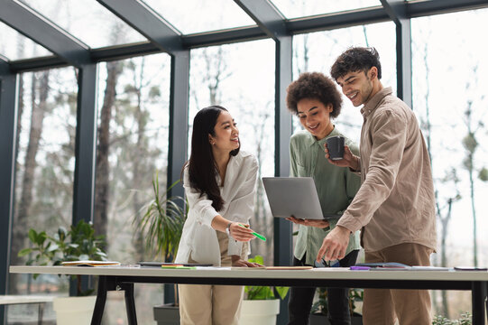 Teamwork. Business Team At Office Interior Working With Laptop And Papers Standing Around Table Together. Three Arabic, African And Asian Colleagues Websurfing Enjoying Professional Cooperation