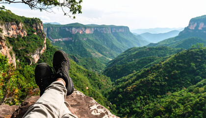 a person sitting on a cliff with a scenic view of mountains and valleys. the person appears to be resting their feet at the edge of the cliff, enjoying the natural surroundings.