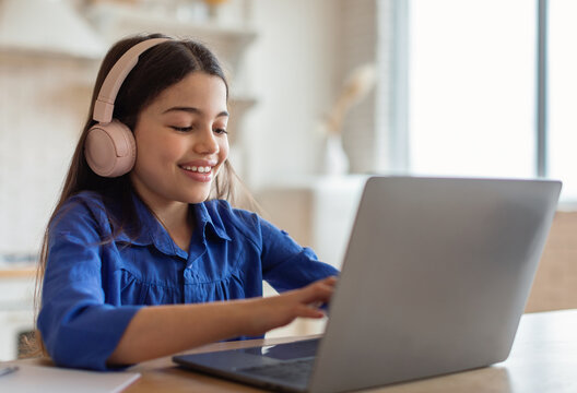 E-Learning Class. Cheerful Arabic Schoolgirl Studying Online Typing On Laptop Computer Wearing Earphones Sitting At Home. School Kid Girl Websurfing Enjoying Distance Lesson